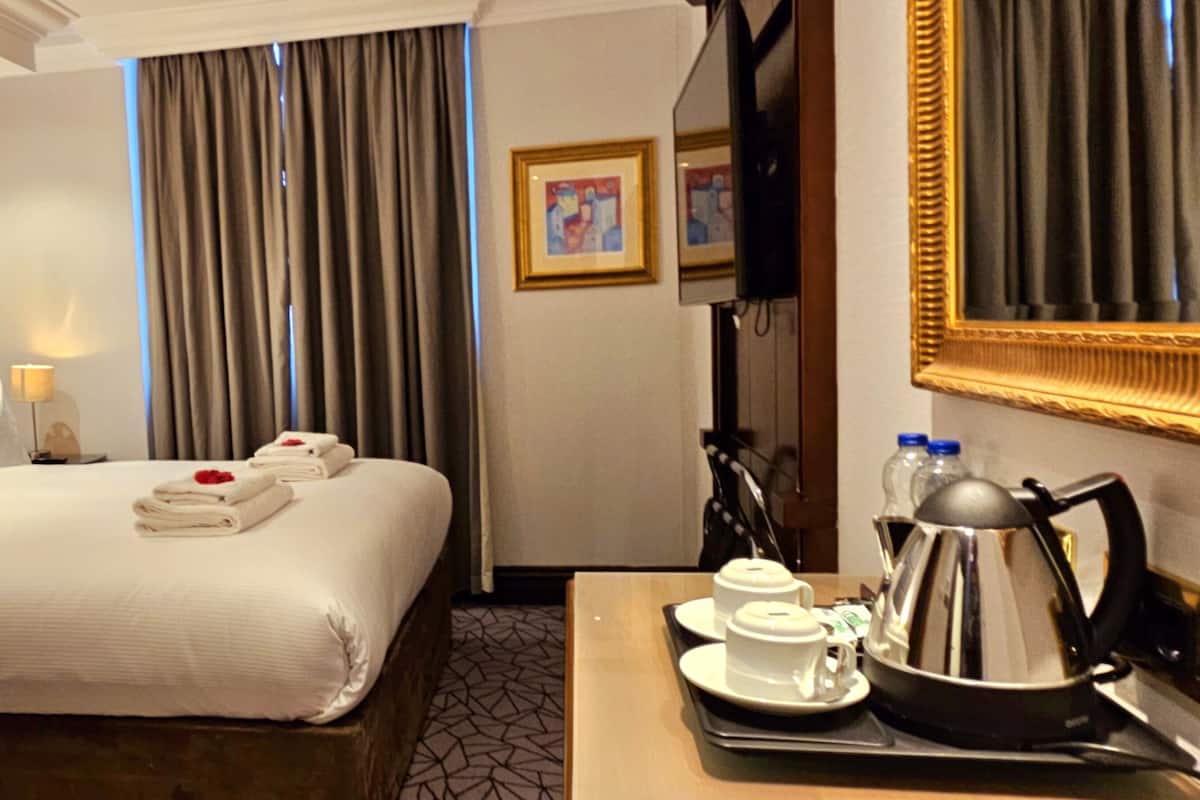 A hotel room with a large bed, a bedside table with a lamp, a coffee maker, and a tray with cups and a teapot.