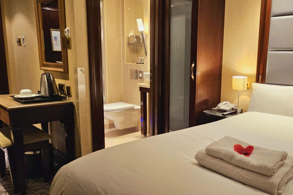 A hotel room with a bed, a bedside table with a lamp, a telephone, and a bathroom visible through an open door.