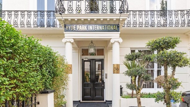 La entrada al Hyde Park International, con una puerta negra, columnas blancas y un balcón con barandilla de hierro forjado.