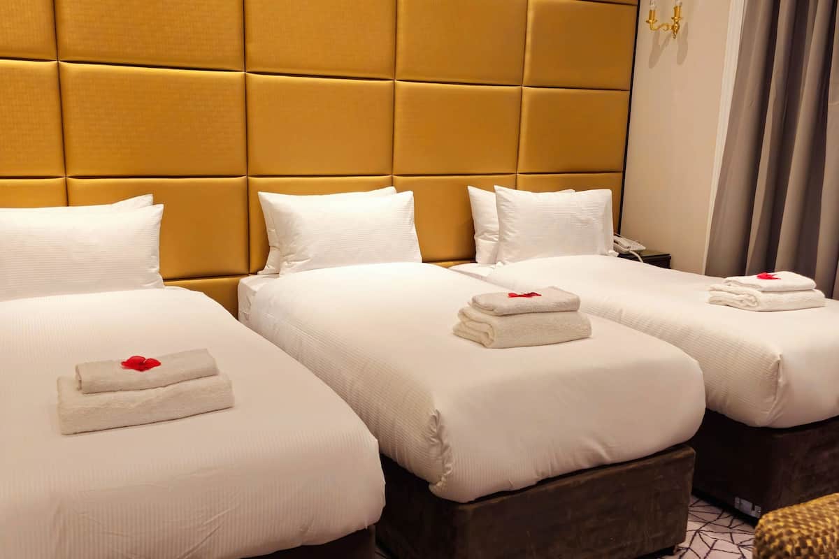 A hotel room with three beds, each with white linens and a red rose on the pillows. There is a nightstand with a lamp and a phone on it. The room has a modern design with a large headboard and a curtain on the window.