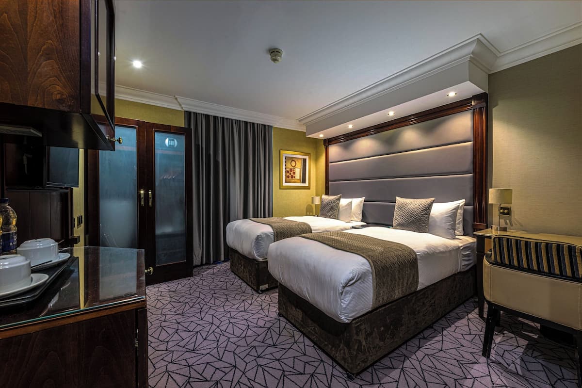 A hotel room with a large bed, two bedside tables, a desk with a chair, a mirror, and a window with curtains.