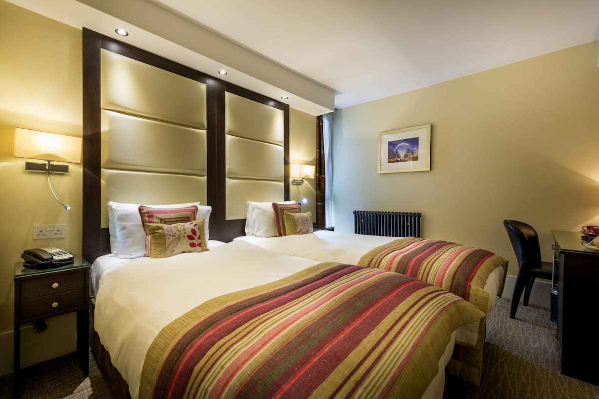 Hotellihuone, jossa on kaksi sänkyä, työpöytä, tuoli, lamppu ja taulu seinällä.