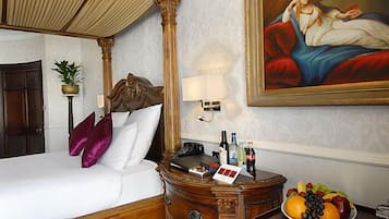 Une chambre d’hôtel avec un lit à baldaquin, une table de chevet sur laquelle se trouve un panier de fruits, un tableau au mur et une lampe.