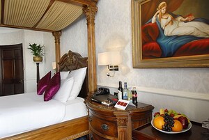 A hotel room with a four-poster bed, a nightstand with a fruit bowl, a painting on the wall, and a lamp.
