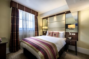 A hotel room with a large bed, a bedside table with a lamp, a mirror, and a window with curtains.