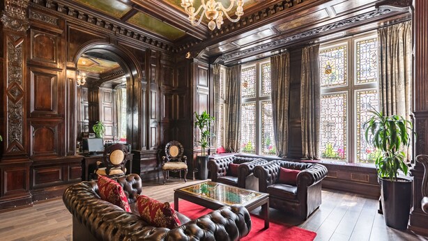 Une chambre luxueuse avec des boiseries sombres, des canapés en cuir, un tapis rouge et de grandes fenêtres ornées de vitraux.