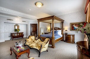 A four-poster canopy bed with a blue bedspread, a chaise lounge, a wooden dresser, and a mirror with a floral arrangement.
