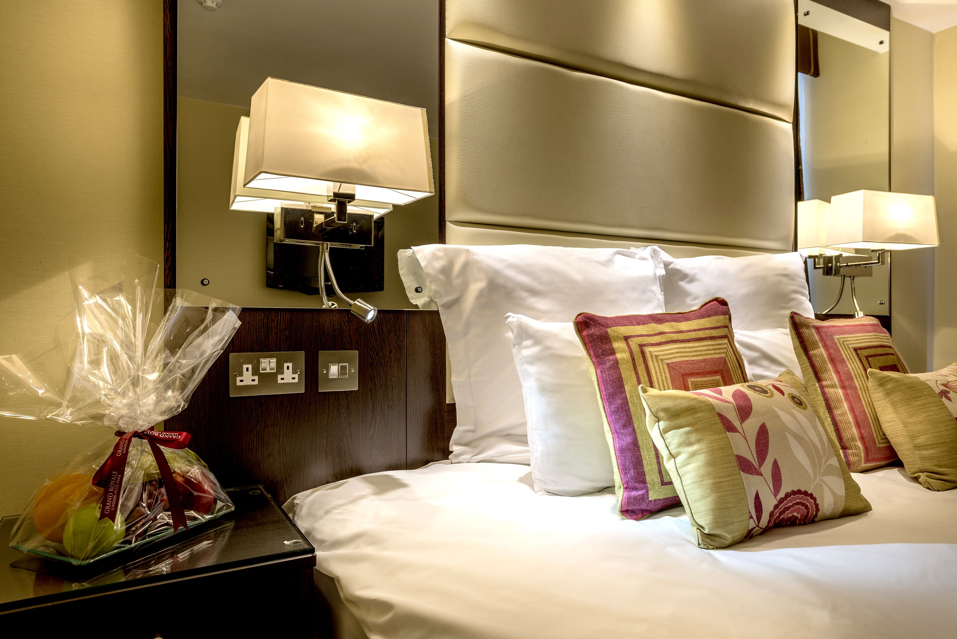 A hotel room with a neatly made bed, a bedside lamp, a nightstand with a lamp, and a wall-mounted control panel.