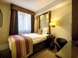 Quarto de hotel com cama, mesa de cabeceira, abajur e secretária com cadeira.