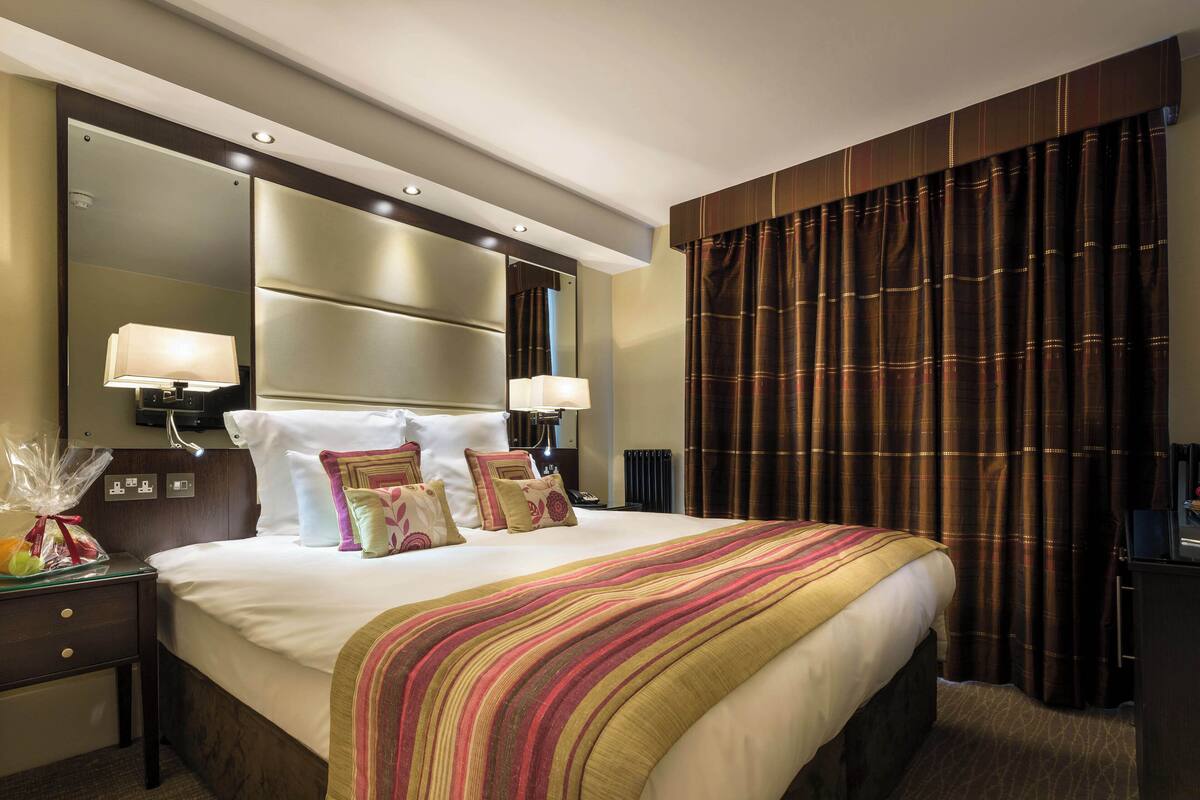 Hotellihuone, jossa on suuri sänky, kaksi yöpöytävalaisinta, yöpöytä, peili ja tumma verho.