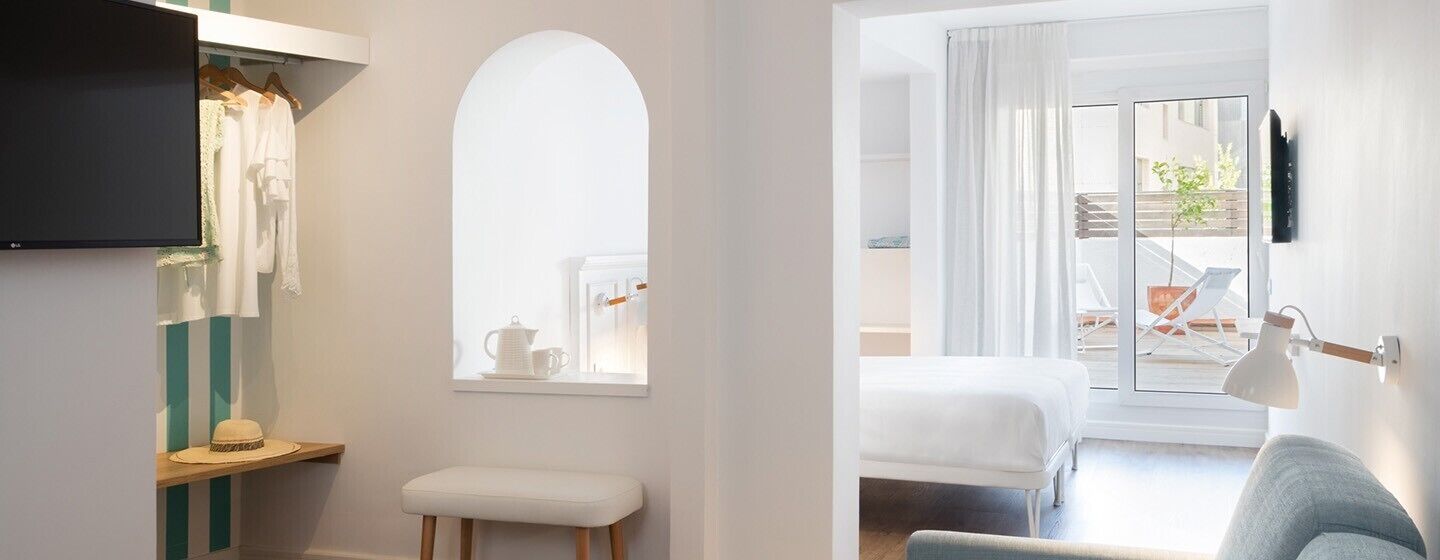 A room with a bed, a bench, a TV, a window with curtains, and a view of the outside.