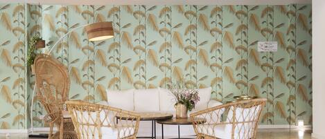 A room with a green patterned wallpaper, a wicker sofa, two wicker chairs, a small table with a vase, and a floor lamp.