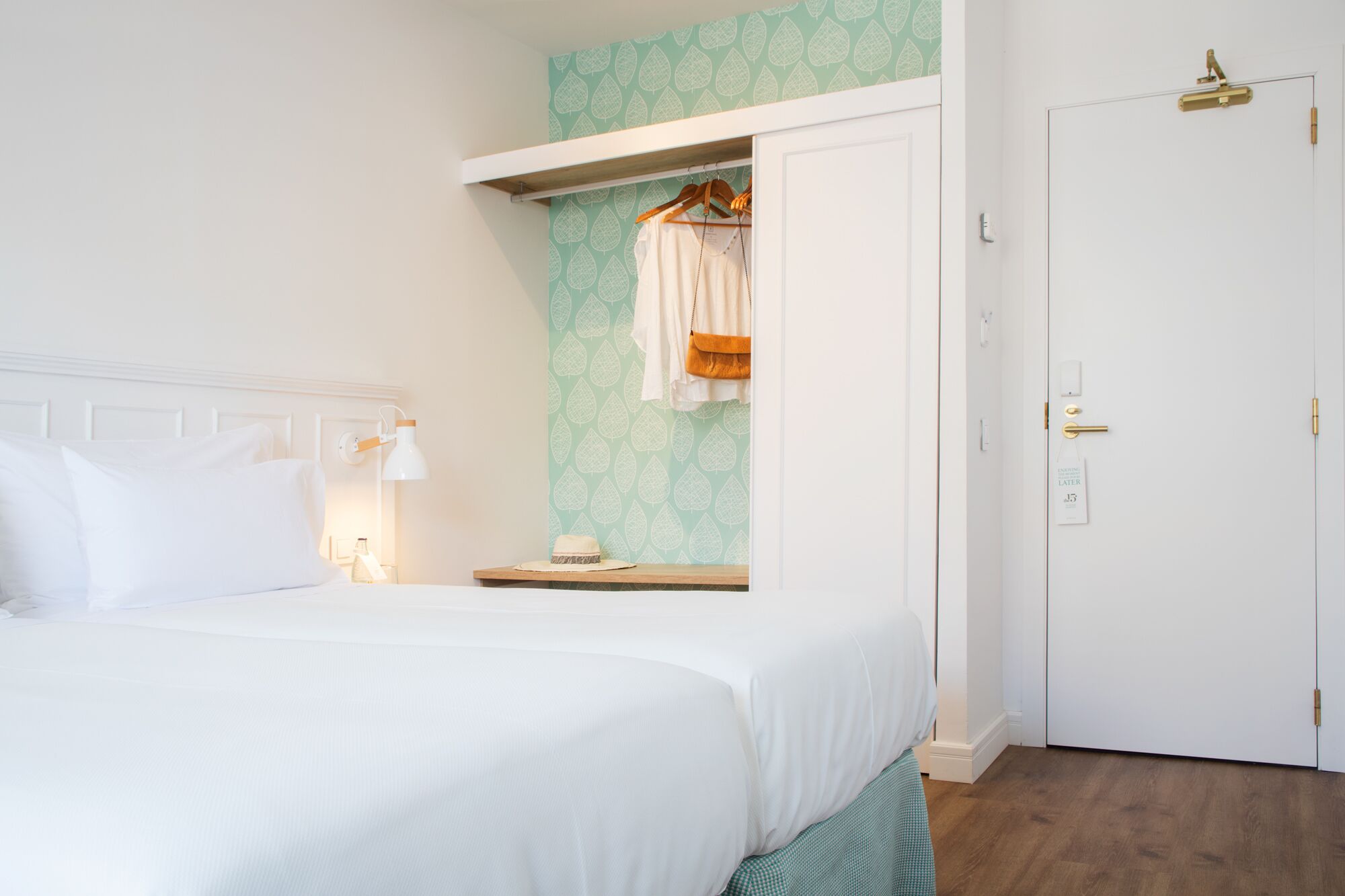 A neatly made bed in a hotel room with a white headboard, a hanging coat rack with a white shirt and a brown bag, a bedside table with a lamp, and a door with a gold handle.