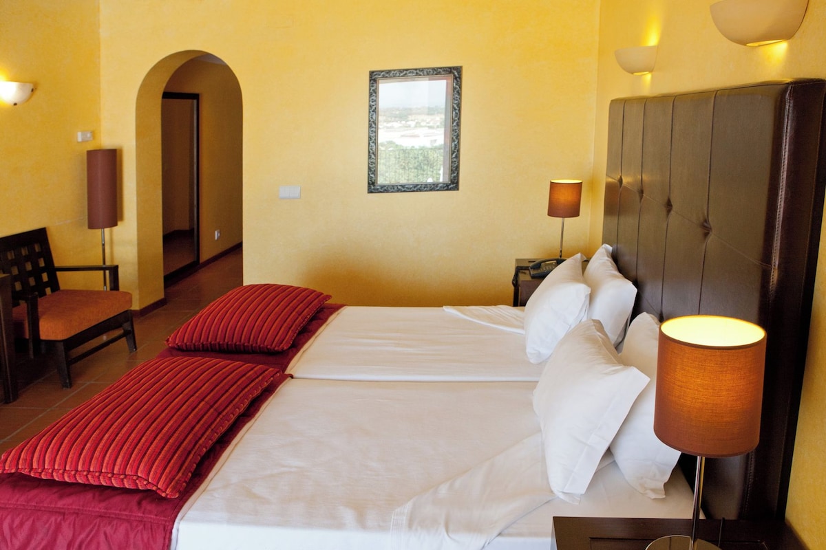 A hotel room with a bed, two pillows, a lamp, a chair, and a framed picture on the wall.