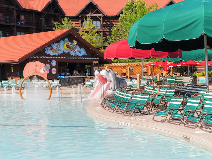 A pool area with lounge chairs and umbrellas, a wooden building with a bear mural, and a log cabin structure in the background.