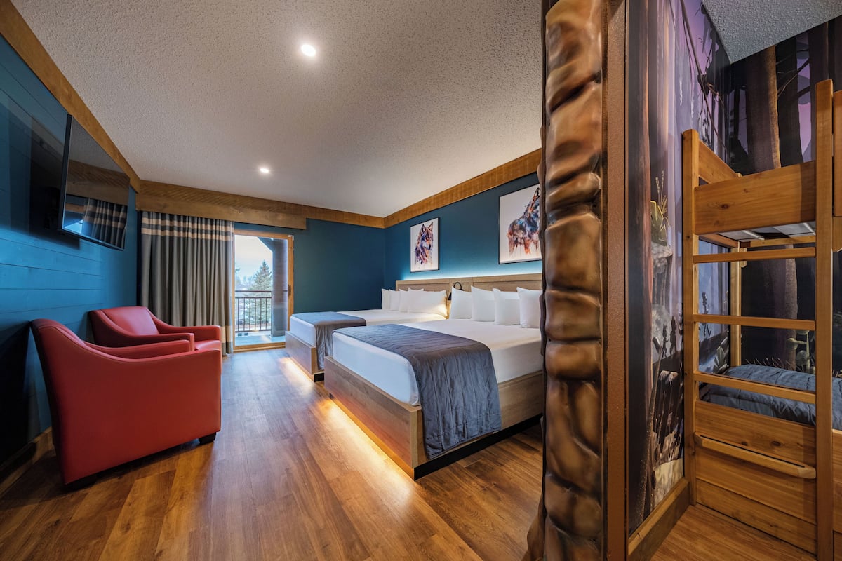 A hotel room with a bed, two red chairs, a wooden bunk bed, and a large window.