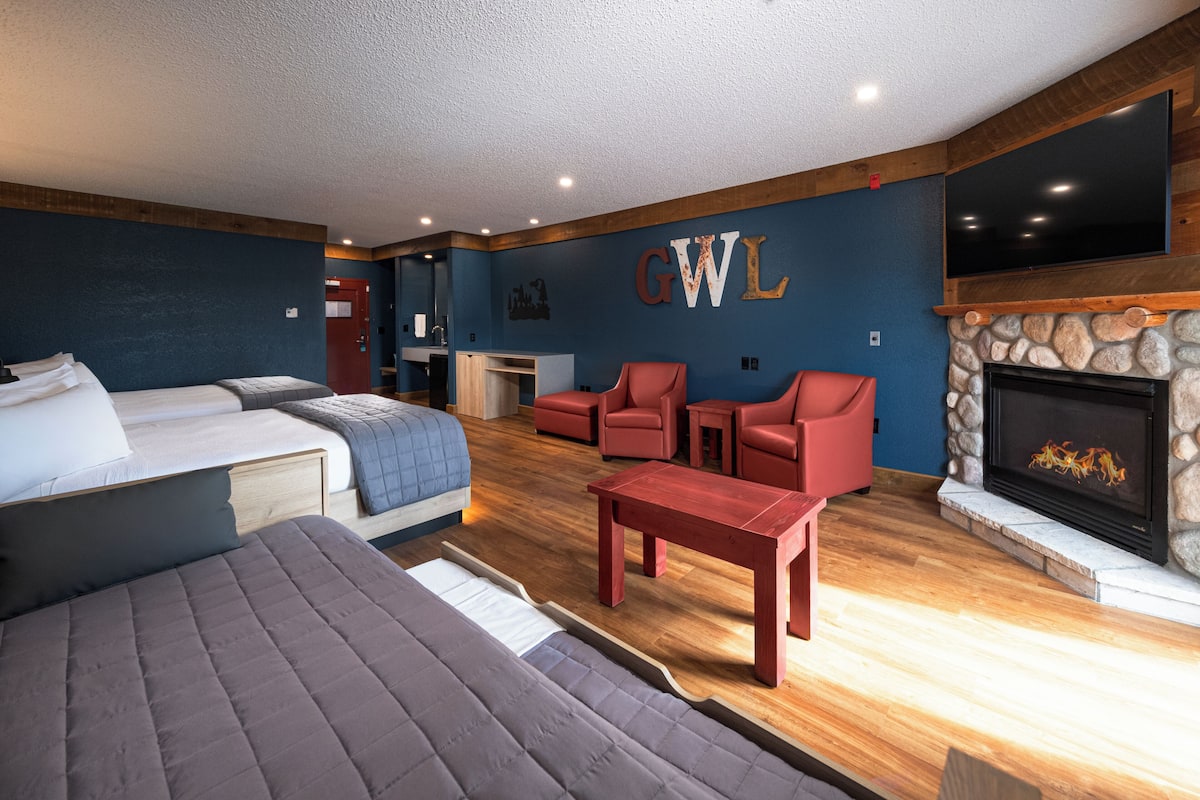 A modern hotel room with a bed, a sofa, a red table, a fireplace, and a TV mounted on the wall.