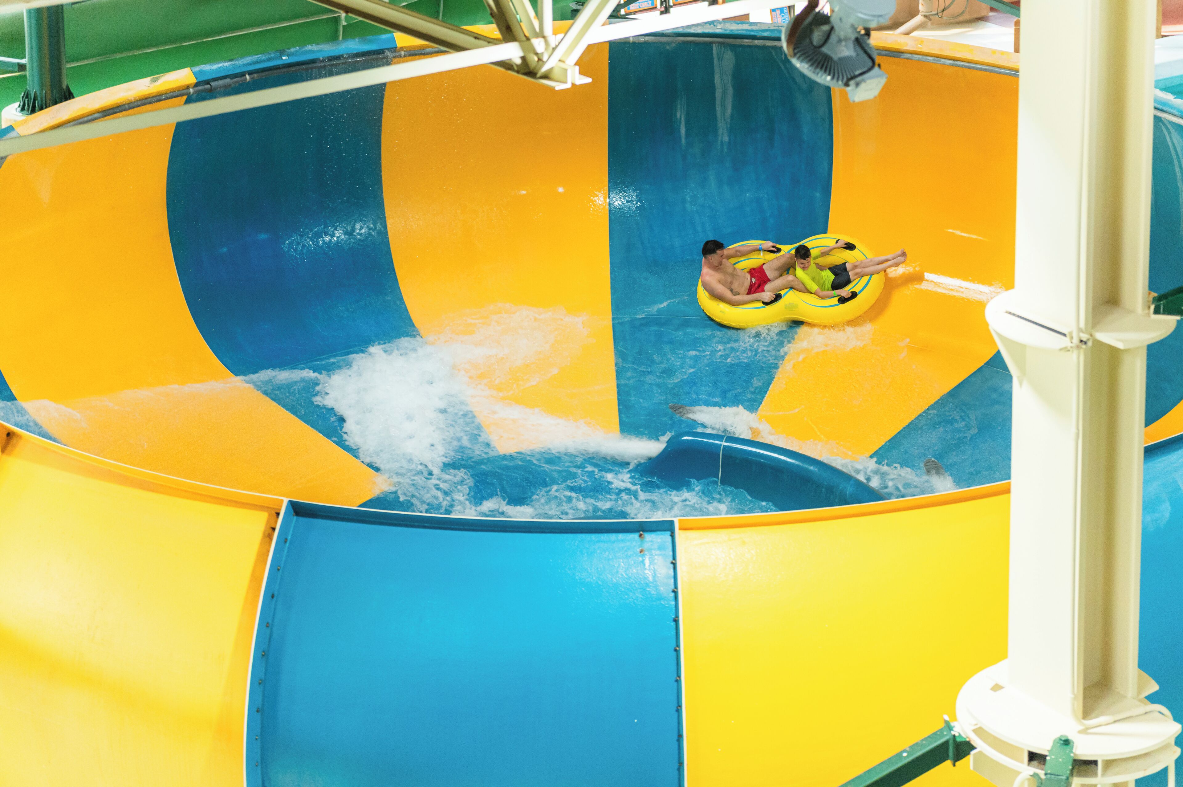A water slide with yellow and blue stripes, a man in a yellow tube, and a person in a red swimsuit.