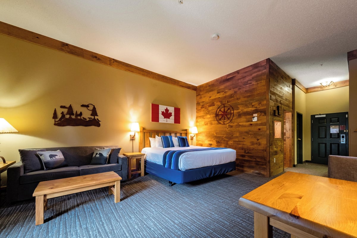 A hotel room with a bed, a sofa, a wooden coffee table, a wall with a Canadian flag, and a wooden wall panel.