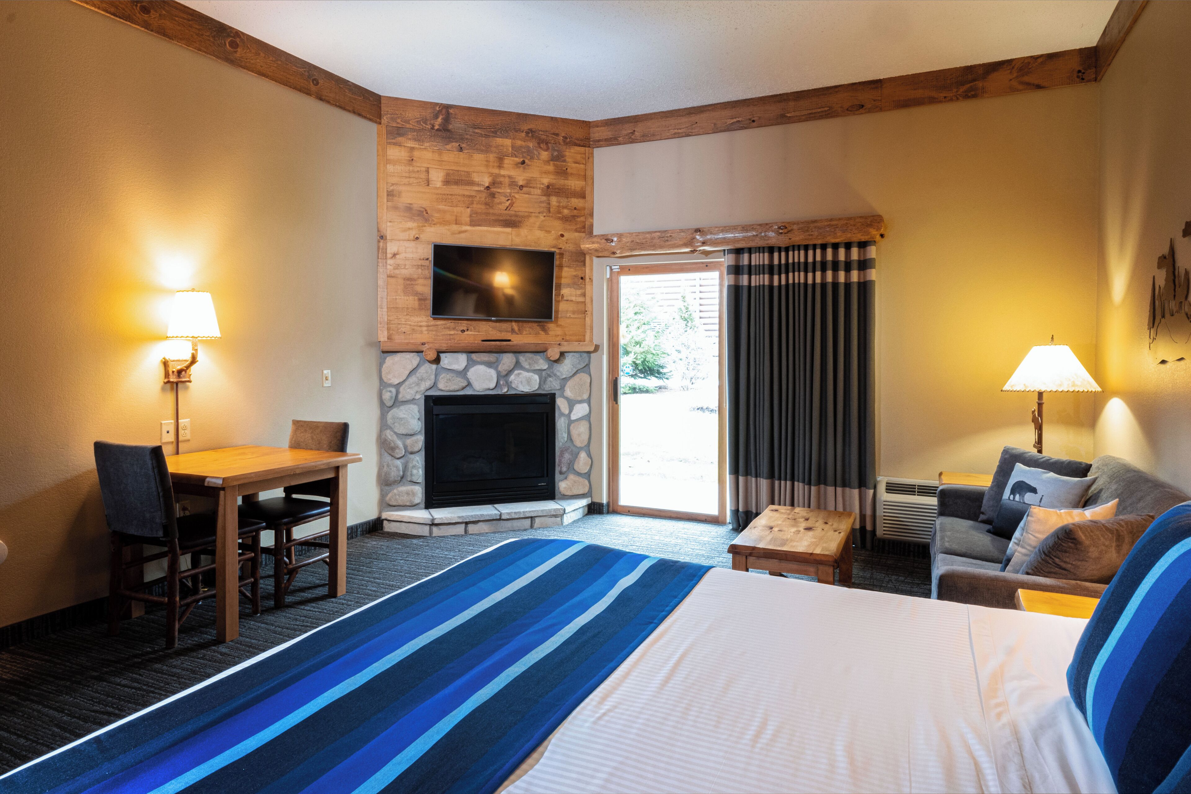 Habitación de hotel con chimenea de piedra, un televisor montado encima, una cama con alfombra a rayas azules, un sofá y un escritorio con una silla.