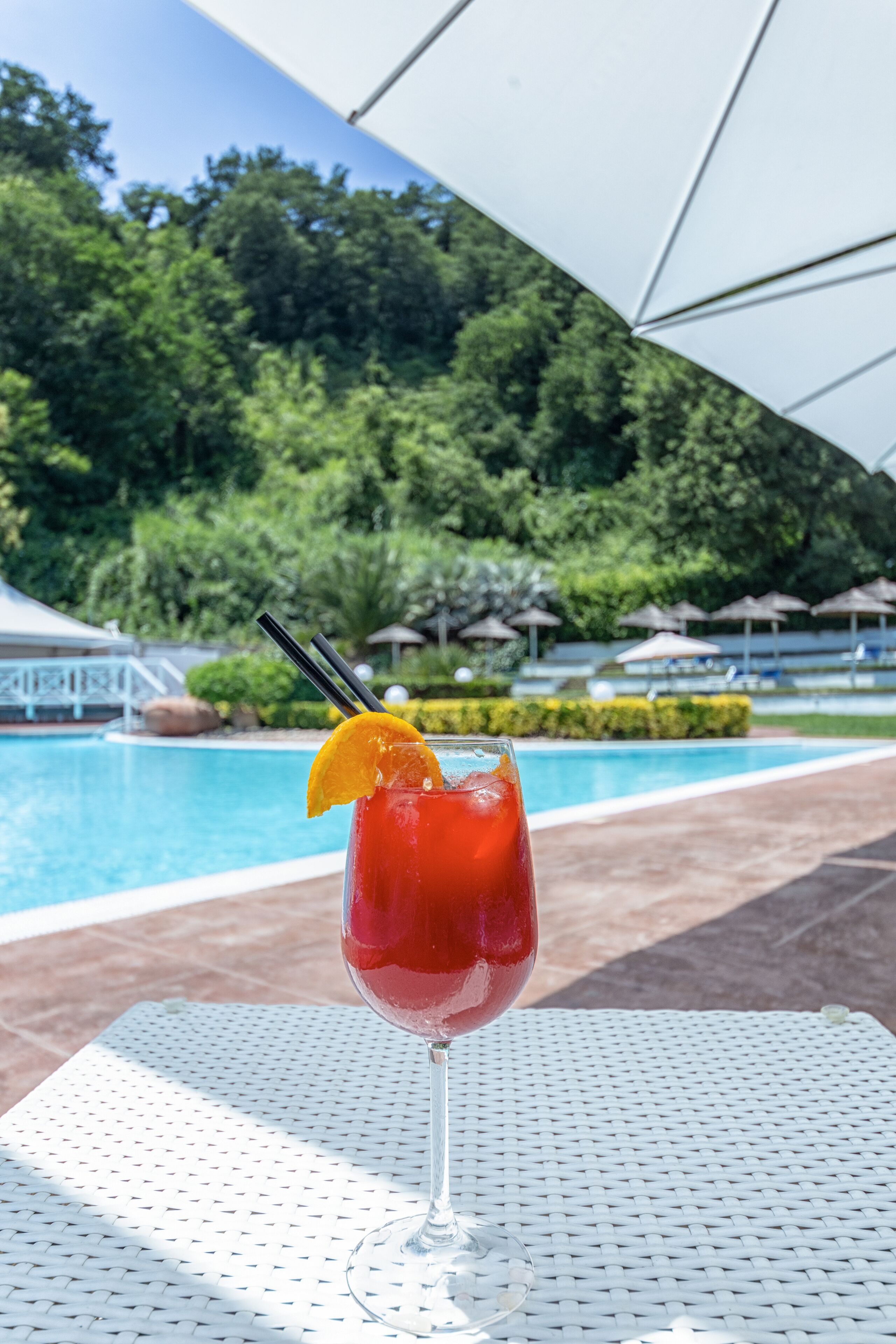 A glass of red drink with a lemon slice and straw on a white table by a pool with umbrellas and greenery in the background.