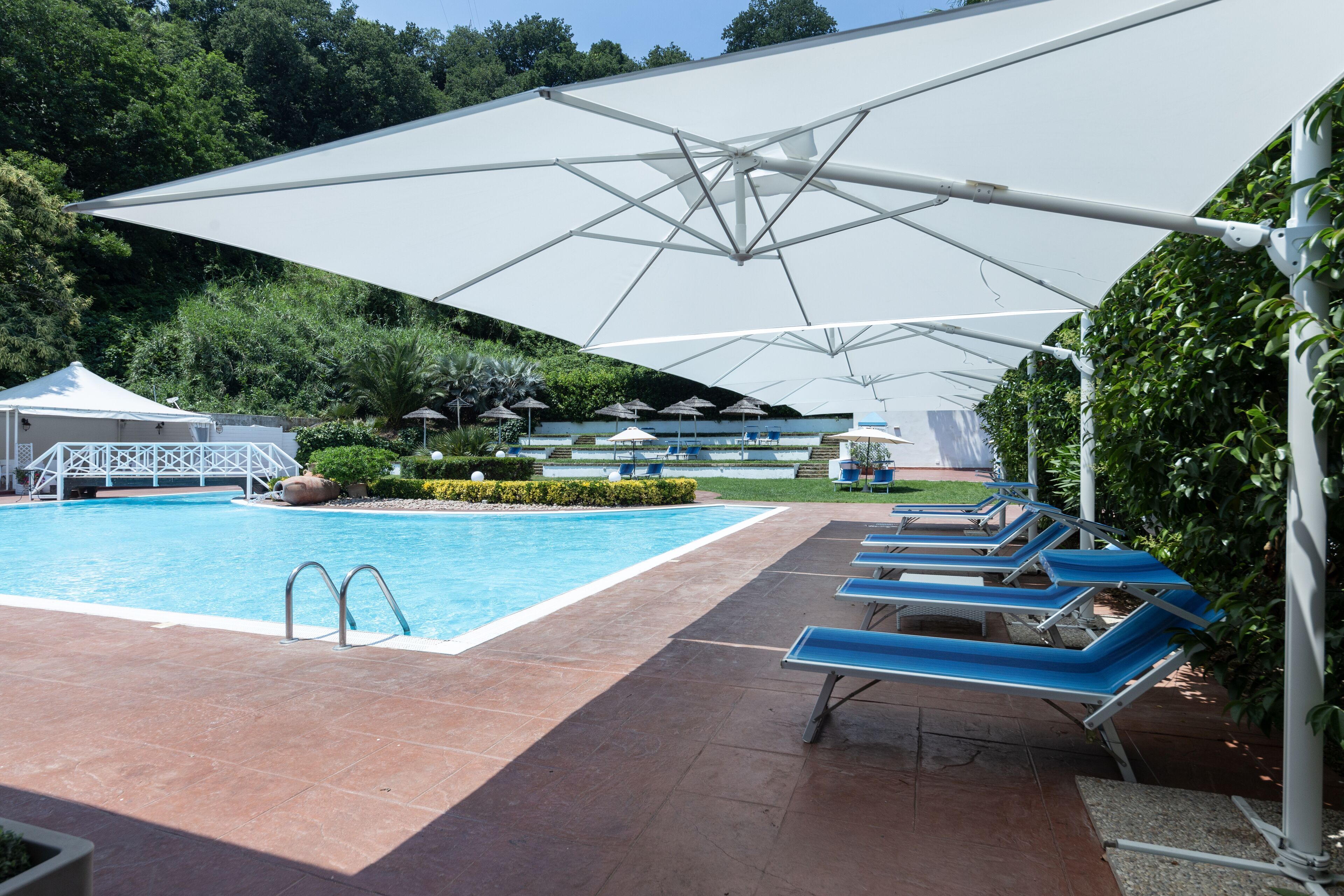 A swimming pool with lounge chairs and umbrellas, surrounded by greenery and a white building.