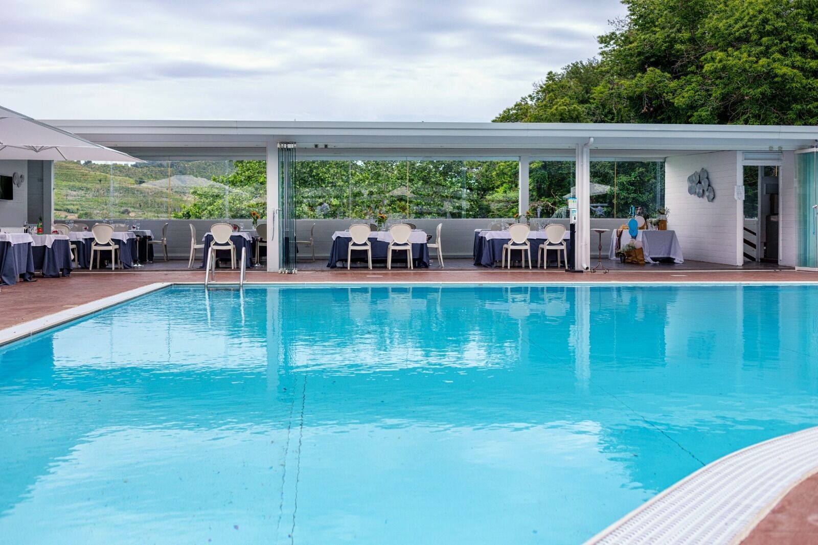 A pool area with tables and chairs under a covered section, surrounded by greenery.