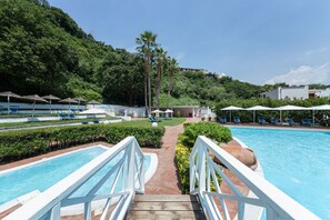 A swimming pool with a wooden deck and white railings, surrounded by lounge chairs and umbrellas, with a lush green hillside in the background.