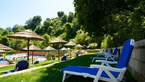 Un área junto a la piscina con sillones de descanso y sombrillas, rodeada de abundante vegetación.