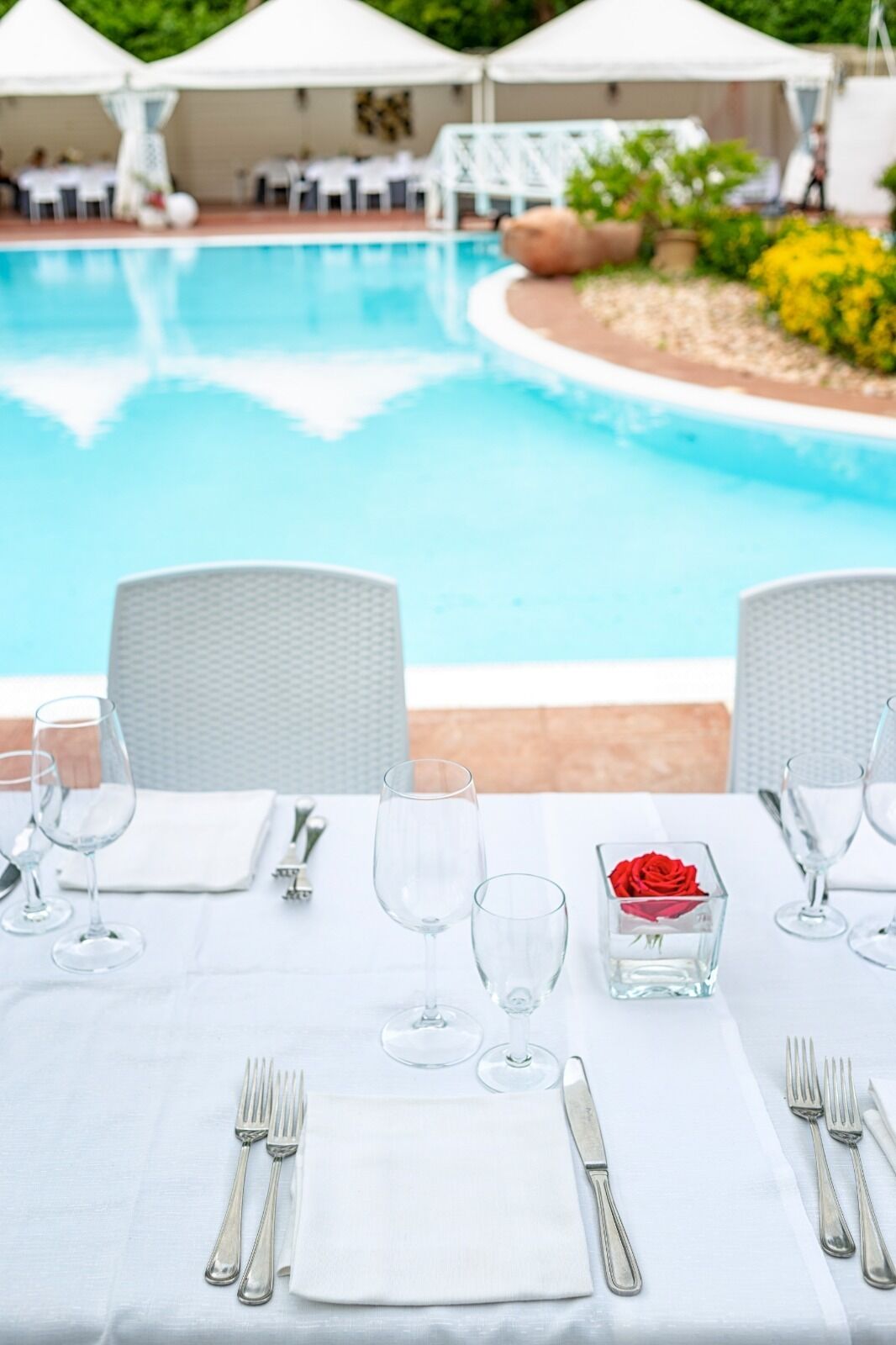A dining table set for four with white tablecloths, silverware, and wine glasses, overlooking a swimming pool with white umbrellas and lounge chairs.