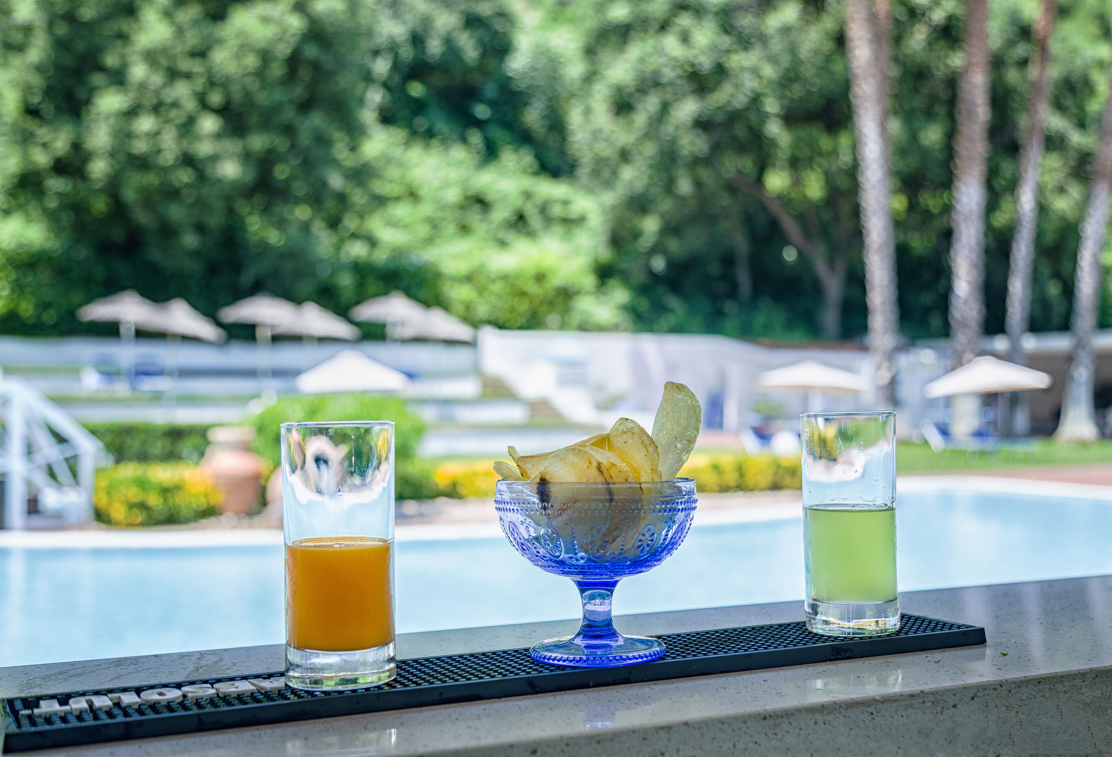 Un espace bar avec vue sur la piscine, comprenant un grand saladier en verre bleu rempli de chips et deux verres contenant des boissons.