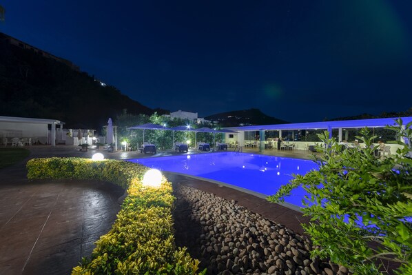 A nighttime view of a swimming pool area with a covered seating area and a mountain backdrop.