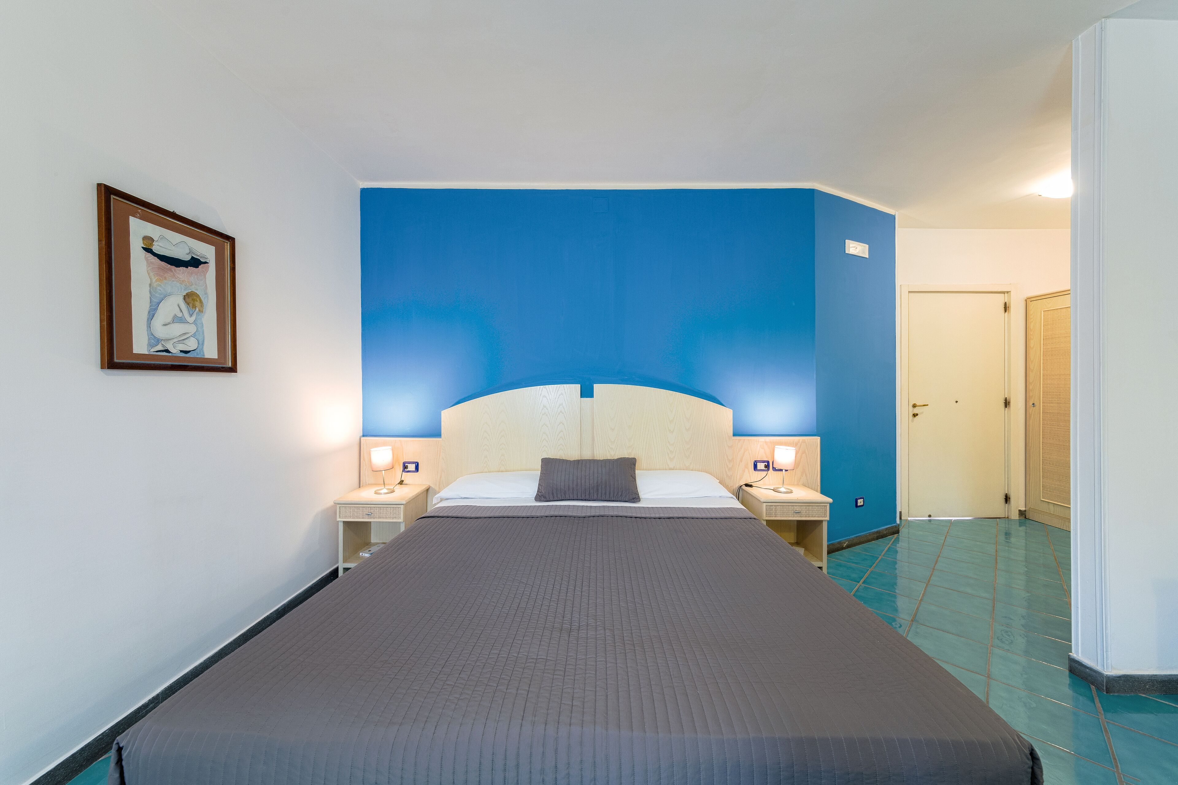 A hotel room with a large bed, two bedside tables with lamps, a framed artwork on the wall, and a blue accent wall.