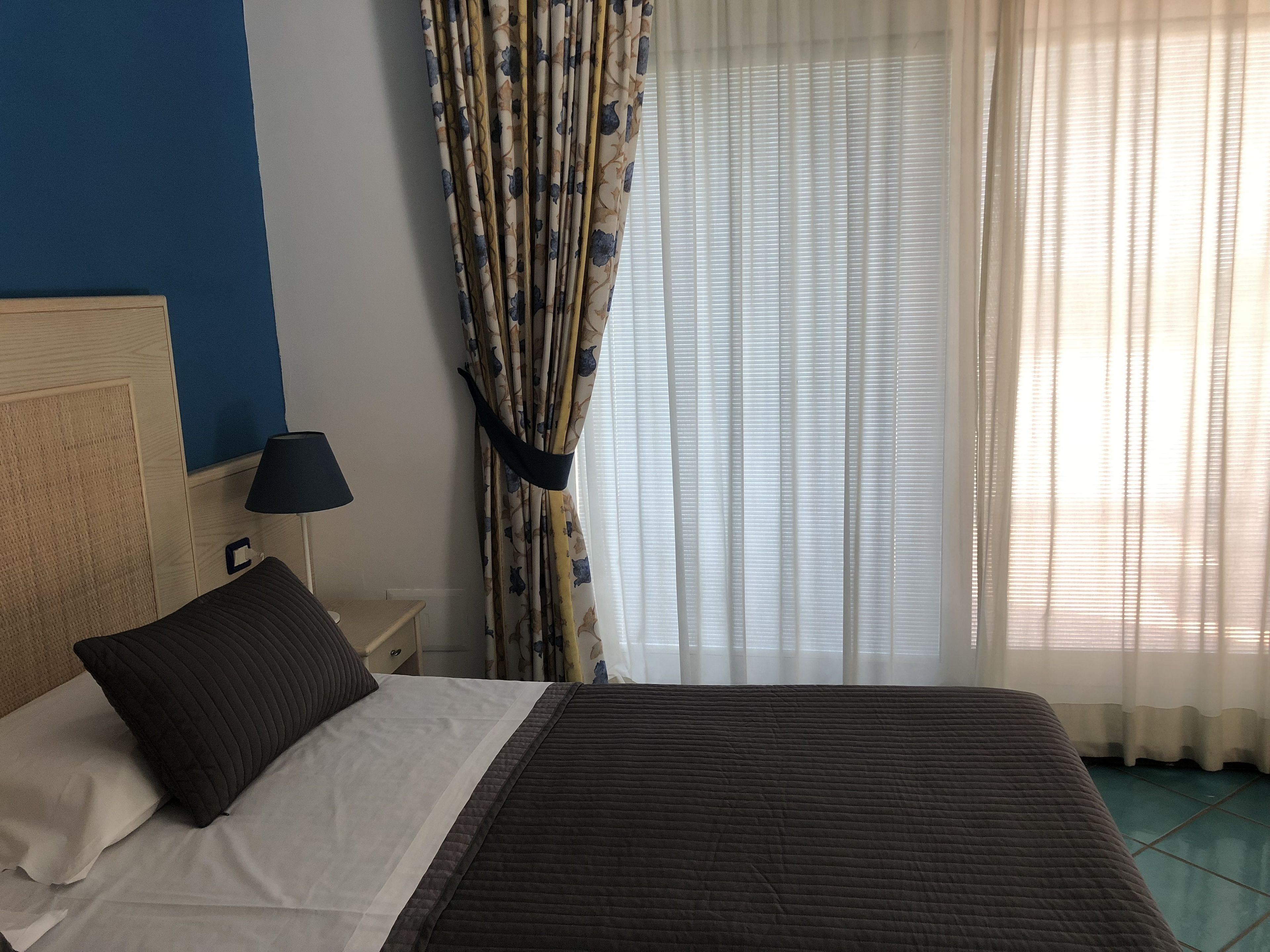 A hotel room with a bed, a nightstand, a lamp, a window with curtains, and a blue wall.
