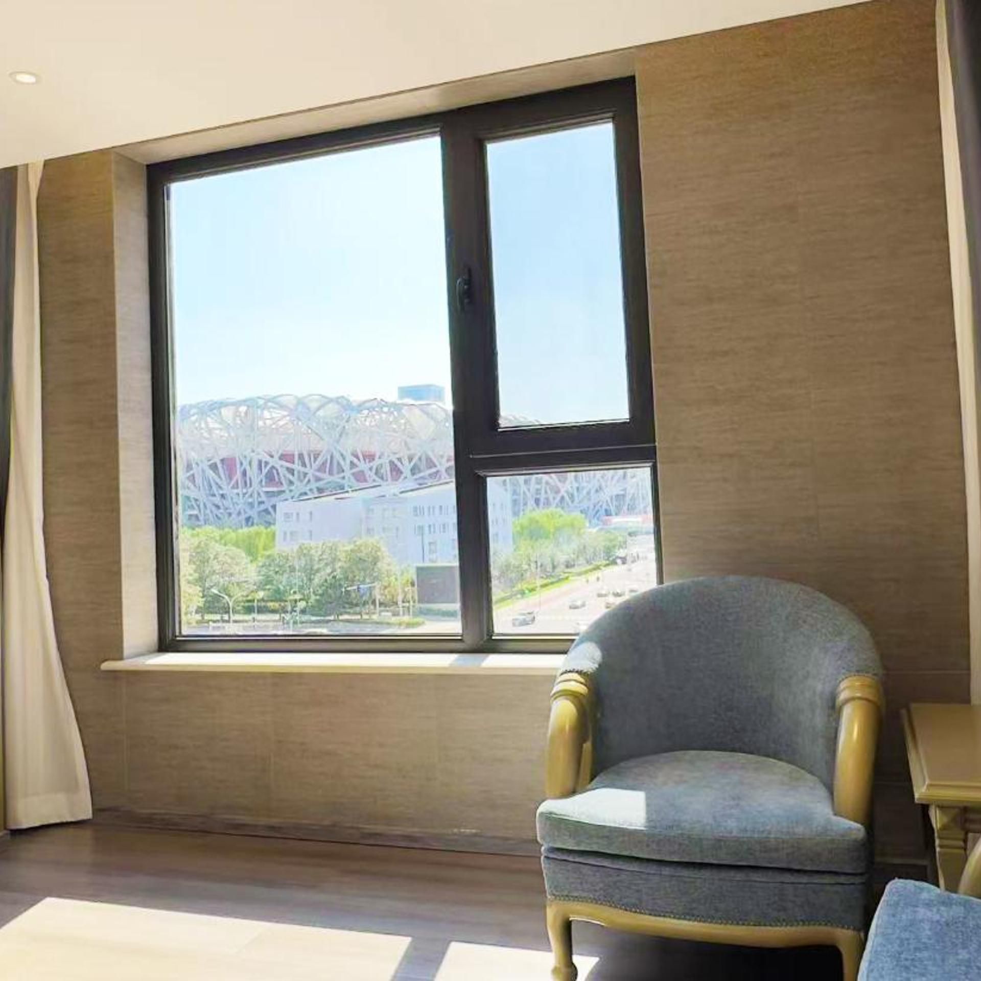 Quarto com uma janela grande, uma cadeira e vista para um edifício com estrutura singular.