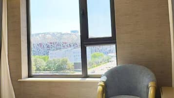 Quarto com uma janela grande, uma cadeira e vista para um edifício com estrutura singular.