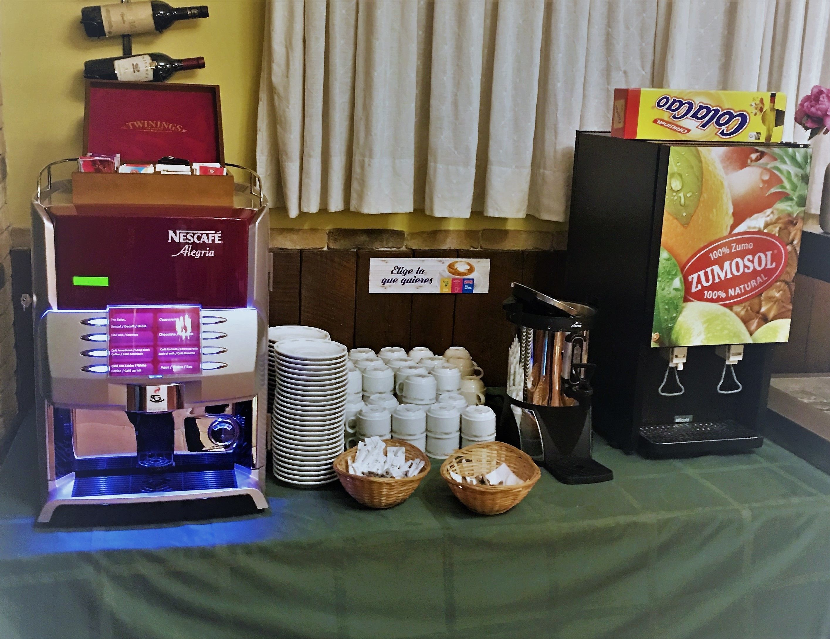 Una estación de café con una máquina Nescafé Dolce Gusto, pilas de vasos desechables y una caja de Zumo Zucchini.