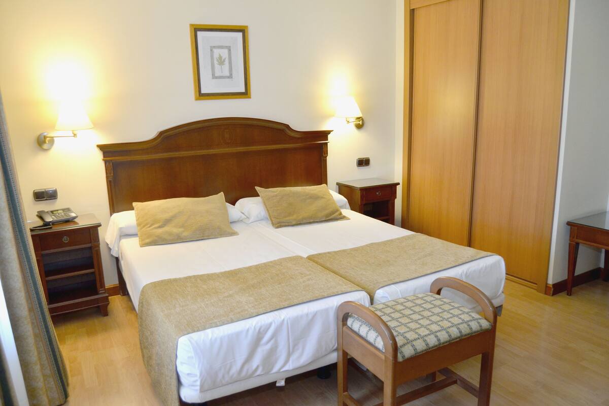 A double bed with two pillows, a bedside table with a lamp, and a wooden chair with a cushion.