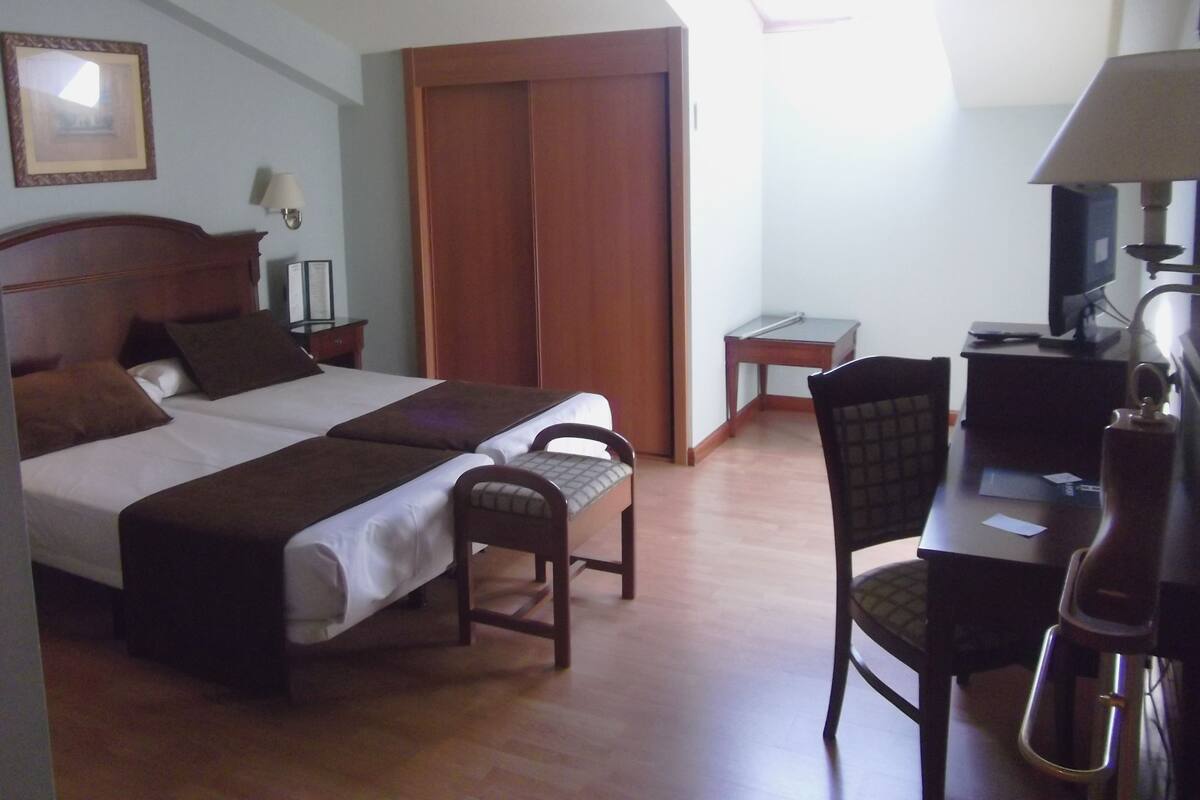 A hotel room with two beds, a desk, a chair, a TV, and a lamp.