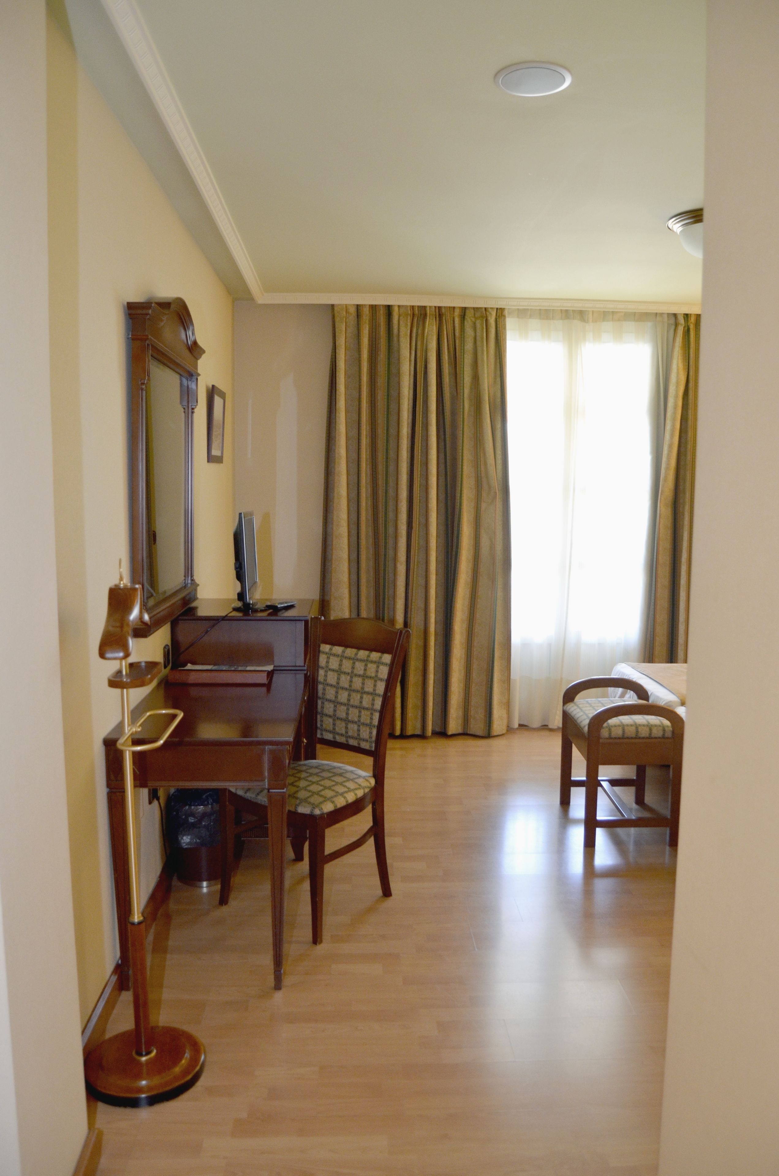 A hotel room with a wooden desk, a chair, a television, and a large window with curtains.