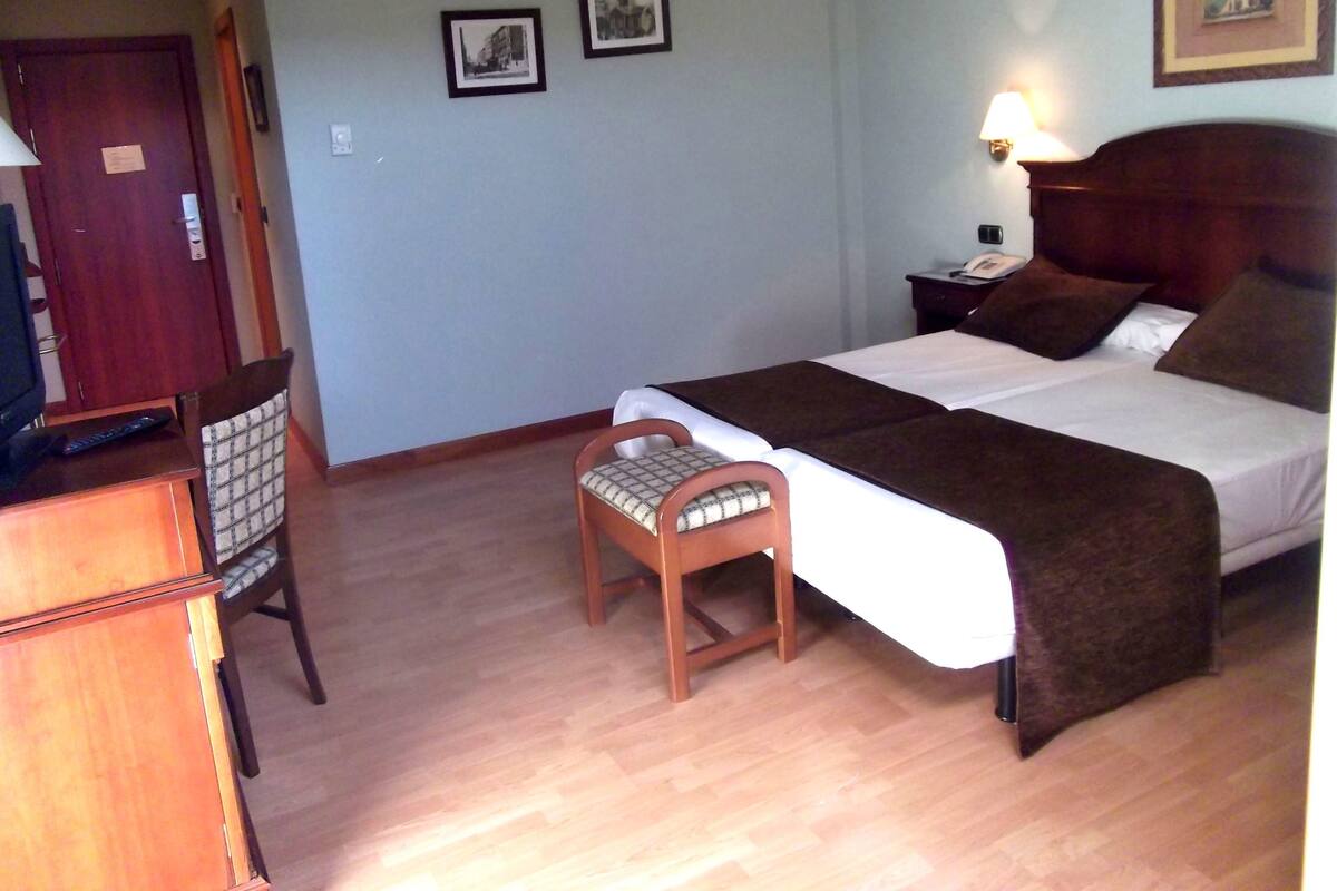A hotel room with a bed, bedside table, lamp, chair, and a desk with a television.