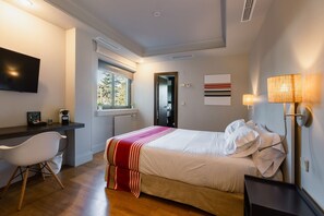 A bedroom with a bed, bedside lamp, desk, chair, window, and artwork on the wall.