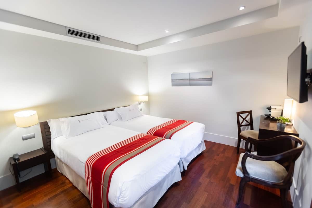 A hotel room with two beds, a desk, a chair, and a painting on the wall.