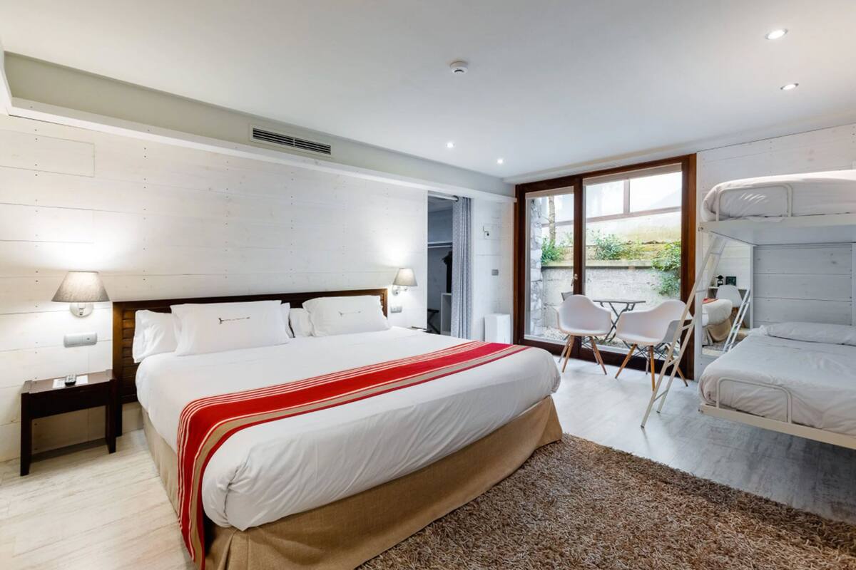 A modern bedroom with a large bed, a small table, two chairs, and a window with a view of greenery.