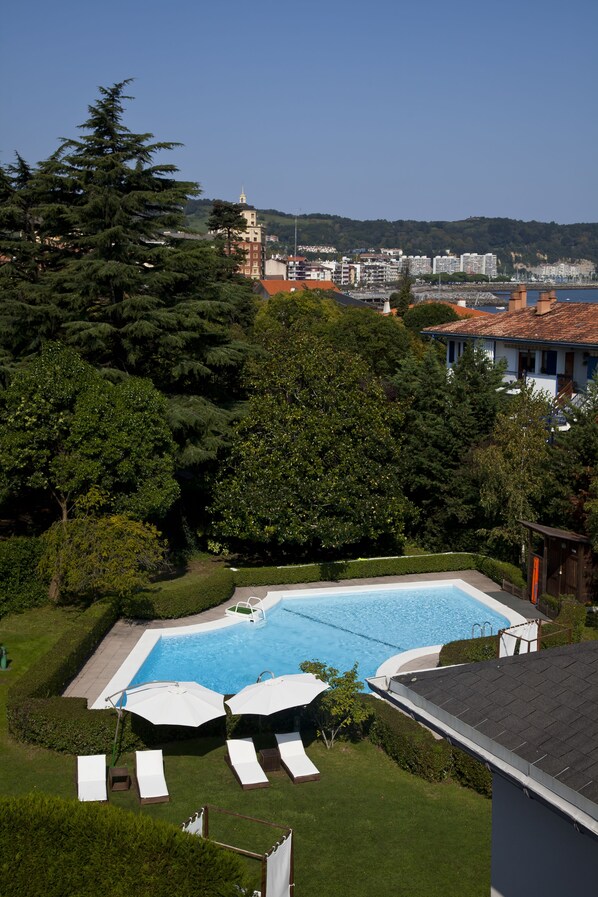 A swimming pool with lounge chairs and umbrellas, surrounded by lush greenery and a view of a cityscape in the background.