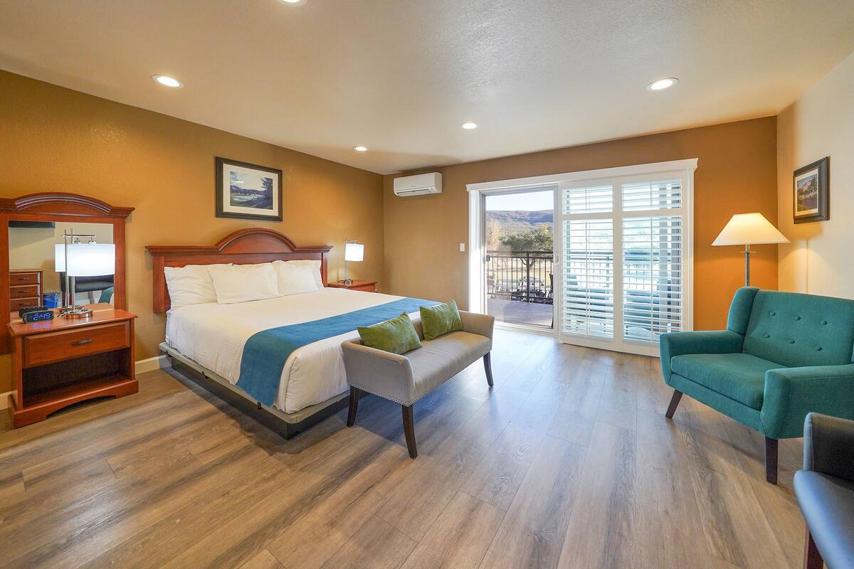 A hotel room with a large bed, a sofa, a chair, a nightstand, a lamp, and a balcony with a view.