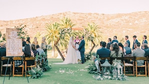A wedding ceremony with guests seated on wooden chairs, a couple at the altar, and a backdrop of palm trees and flowers. - San Vicente Golf Resort (Ramona)