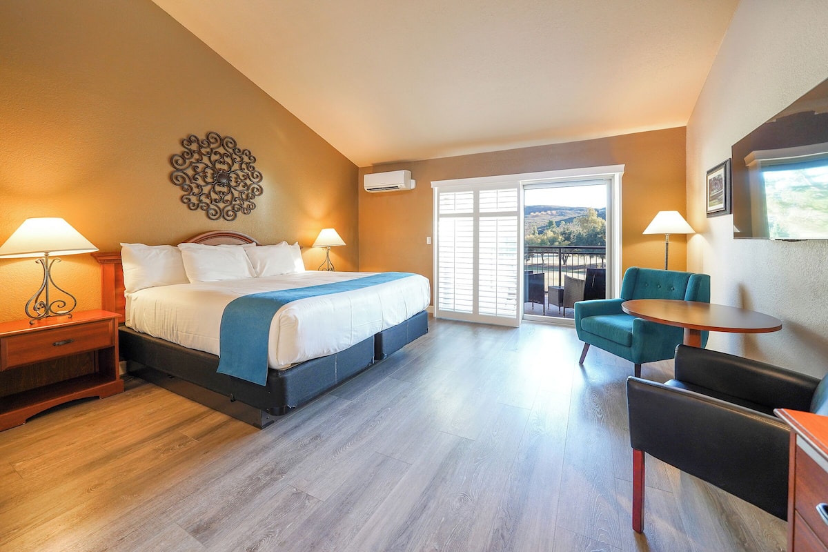 A hotel room with a bed, two bedside tables with lamps, a blue chair, a small table, and a balcony with a view.