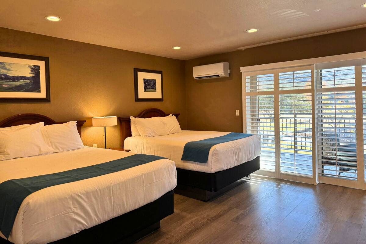 A hotel room with two beds, a large window with blinds, an air conditioning unit, and a framed picture on the wall.