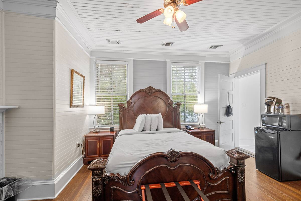 A bedroom with a wooden bed, white walls, a ceiling fan, and a refrigerator.